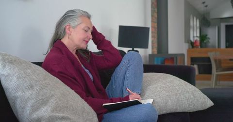 Mature woman writing in notebook on sofa wearing burgundy cardigan, cozy home journaling