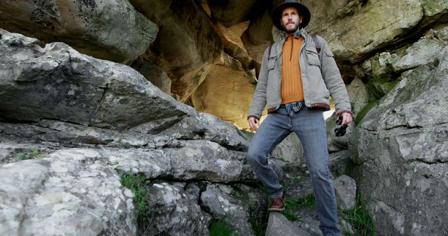 Man in rugged outdoor clothing exploring rocky cave while holding camera. Ideal for themes related to adventure, travel, exploration, nature photography, and outdoor activities. Can be used in travel blogs, adventure magazines, or promotional materials for outdoor gear.