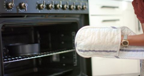 African american woman placing cake pan into oven wearing quilted mitt and wristwatch