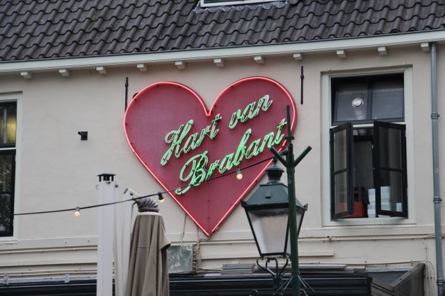 Neon heart-shaped sign displaying 'Hart van Brabant' hanging on building facade. Perfect for illustrating Dutch culture, showcasing vintage typography, or highlighting unique storefronts. Suitable for travel blogs, cultural studies, and architectural features.