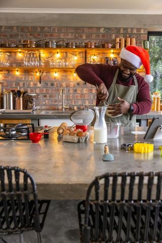 African american man whisking holiday batter in rustic kitchen wearing santa hat
