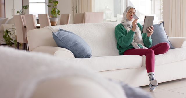 Woman wearing hijab sitting on white couch in modern living room, reading from a tablet, dressed casually in green sweater and striped socks. Living room is bright and cozy with light-colored furniture and decor. Image can be used for topics like technology, modern lifestyle, leisure activities, relaxation, and home interior design.