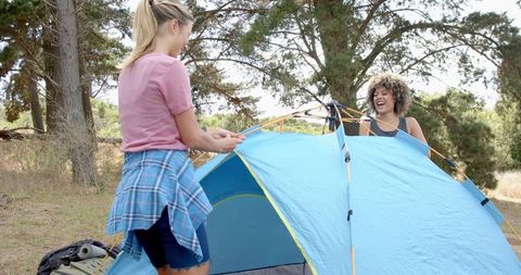 Two women enjoy camping in scenic woodland location setting up tent