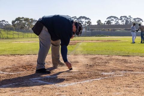 Baseball umpire brushing home plate chalk on sunlit diamond preparing field for play