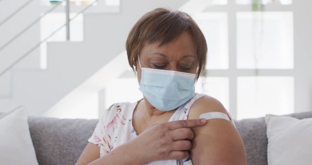 Senior woman wearing face mask showing bandage on her arm, emphasizing vaccination and health safety. Ideal for topics on healthcare, COVID-19 prevention, vaccination efforts among elderly, community health initiatives, and promoting safety during pandemics.