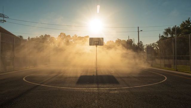 Lonely basketball hoop casting long shadow across misty outdoor court during golden hour