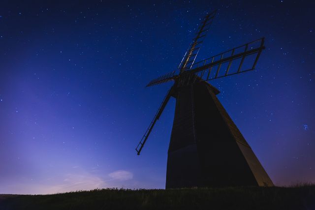 This image captures the majestic silhouette of a historic windmill under a clear, starry night sky. The twilight slowly fading on the horizon adds a touch of calming purple hue, providing a peaceful and tranquil night landscape. Ideal for themes related to nature, historic landmarks, and starry night scenery. Suitable for use in travel blogs, educational content on renewable energy, and relaxation-themed prints.