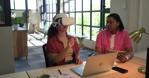African american women testing vr headset and collaborating with laptop in bright coworking office