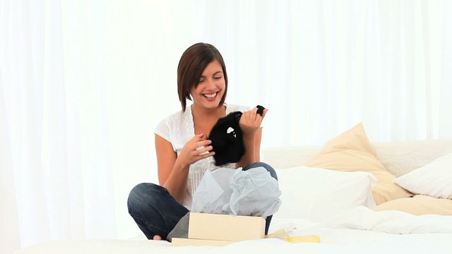 Young woman with short brown hair sitting casually on floor in bright living room. Her expression is one of joyful surprise as she unwraps a gift box containing a thoughtful present. Scene suggests themes of celebration, special occasions, friendship, or loved ones. Ideal for marketing around holidays, events or product launches. Demostrates emotion and modern, cozy home environment.