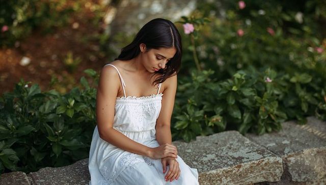 Sitting asian woman in white embroidered sundress clasping hands on stone ledge