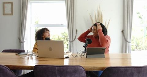 Female colleagues recording podcast at home dining table with laptop and tablet
