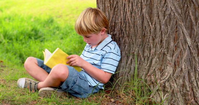 Young blonde boy sitting against large tree trunk in grassy area, engrossed in reading yellow book. Ideal for educational content, children's literature, outdoor learning, and childhood development themes.