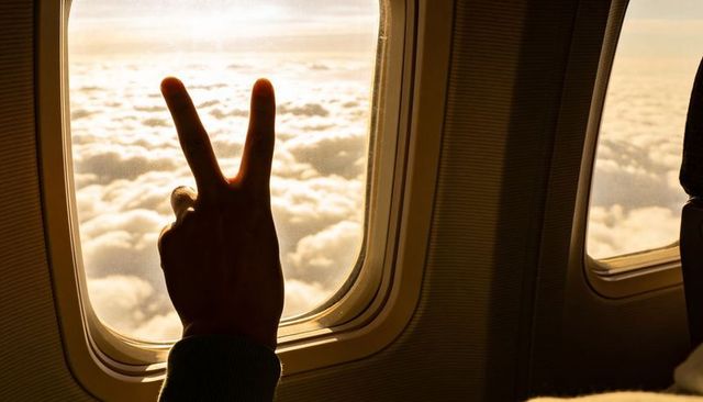Silhouetted hand making peace v gesture at airplane window over golden sunrise clouds