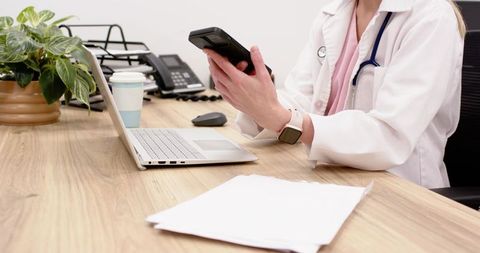 Doctor checking smartphone while reviewing medical documents at clinic desk with laptop