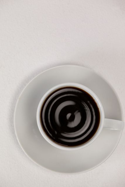 This image shows an overhead view of a cup of black coffee on a white saucer against a white background. Ideal for use in articles or advertisements related to coffee, cafes, morning routines, or minimalist lifestyle. Perfect for blogs, social media posts, or websites focusing on beverages, relaxation, or simple pleasures.