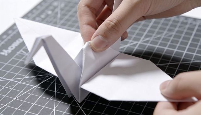 Hands folding origami crane on grid cutting mat, pinching central fold in craft workspace