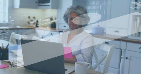 Senior woman working on laptop in kitchen holding pink sticky note with coffee and glasses