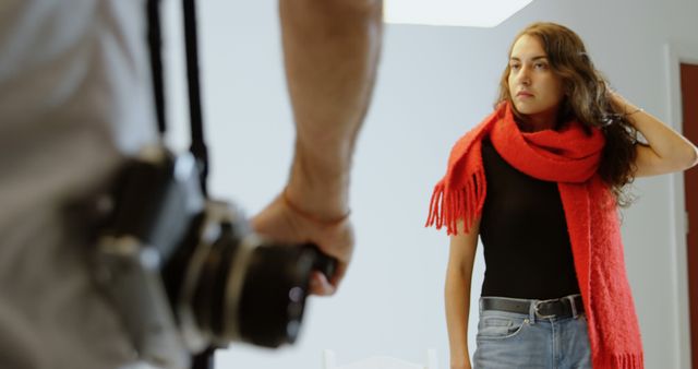 A young Caucasian woman poses for a photographer, adjusting her bright red scarf, with copy space. Her focused expression and the photographer's perspective suggest a professional photoshoot in progress.