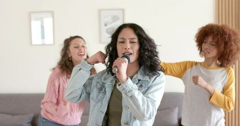 Diverse female friends singing and dancing in cozy living room with wired microphone