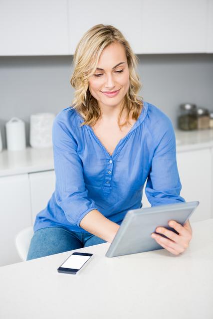Blonde woman sitting in modern kitchen using digital tablet, smiling and relaxed. Ideal for lifestyle blogs, technology advertisements, home decor websites, and articles about modern living and connectivity.