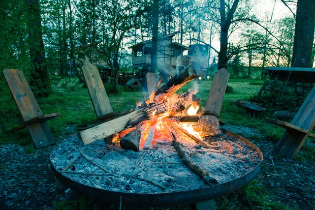Warm campfire with wooden chairs in serene forest clearing at dusk, great for showcasing camping, adventuring, and outdoor relaxation. Suitable for promoting outdoor activities, travel, and nature retreats.