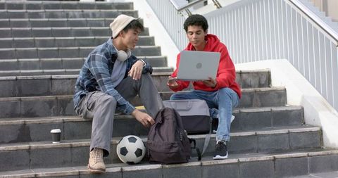 Multicultural male students collaborating on laptop outdoors on campus steps with soccer ball