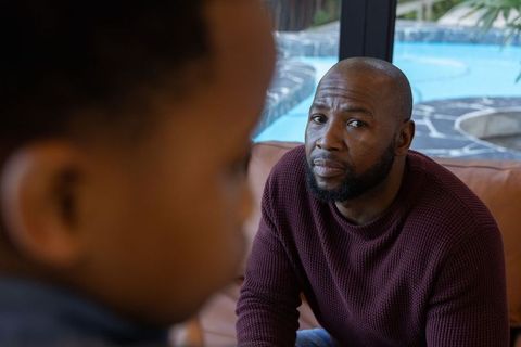 African american father sitting on sofa watching child near patio pool through glass door
