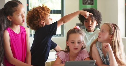 Curious classroom children gathering around tablet, pointing and collaborating on screen