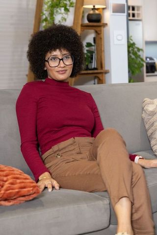African american woman sitting on sofa wearing burgundy top and tan trousers in cozy home