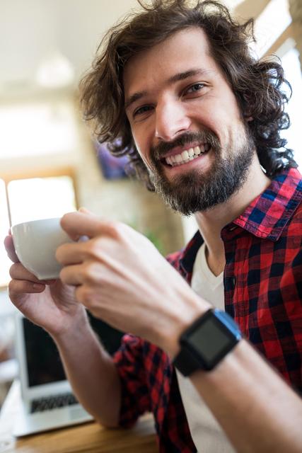 This image shows a young man with a beard smiling while holding a cup of coffee in a cafe. He is wearing a red plaid shirt and a smartwatch, with a laptop in the background. Ideal for use in lifestyle blogs, coffee shop promotions, technology and lifestyle articles, and advertisements focusing on casual and relaxed environments.