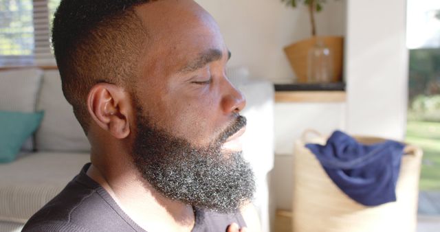 Man with a beard sitting indoors, eyes closed in peaceful meditation. Light illuminates his face, suggesting early morning or late afternoon sun. Perfect for content related to meditation, mental health, self-care, and wellness tips.