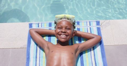 This image depicts a joyful boy relaxing by the edge of a pool. He is wearing swimming goggles and lying on a striped towel, radiating happiness likely due to summer activities and leisure time. This image can be useful for promotional materials related to family vacations, children's swimming lessons, or summer camp advertisements. It captures the essence of enjoyment and carefree moments typically associated with the summer months and waterside adventures.