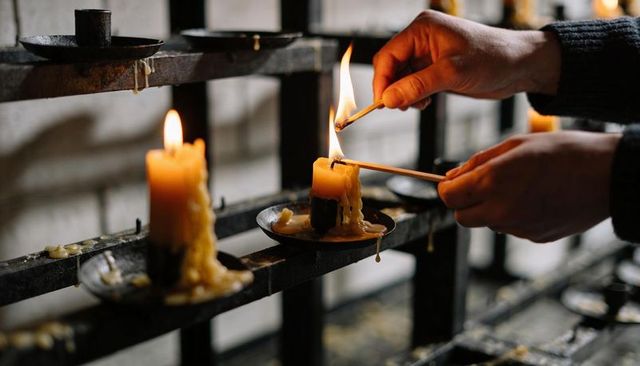 Lighting votive candle on iron rack with melting wax and warm flame, hands in dark sweater