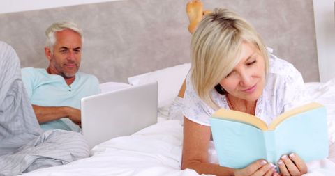 Senior Couple Relaxing with Book and Laptop in Cozy Bedroom