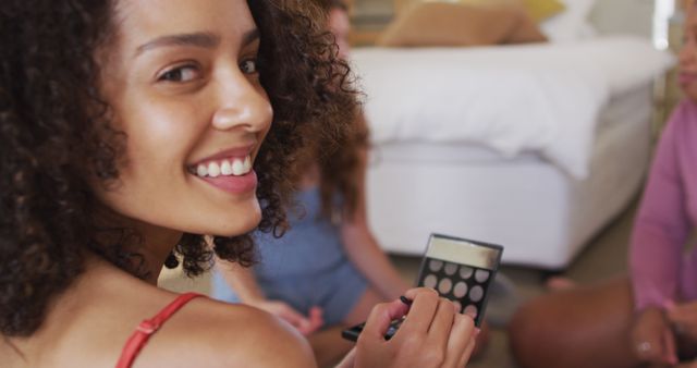 Young woman with curly hair smiling while holding an eyeshadow palette. Scene shows casual home setting, implied conversation with friends. Ideal for use in ads or articles on beauty, makeup tips, self-care routines, and diversity in beauty.