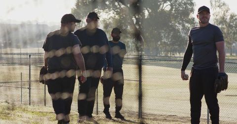 Sunlit baseball players walking with teammate standing holding ball and glove during golden hour