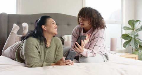 Friends Laughing on Bed Sharing Smartphones in Cozy Pastel Bedroom