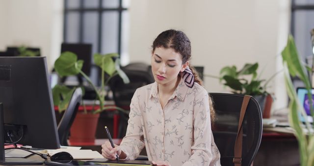 Young Asian businesswoman working at a desk in a modern office. She is notetaking while beside a computer monitor, giving her an appearance of being focused and diligent. Office environment includes potted plants, providing a refreshing and green workspace. Ideal for depicting modern corporate workspaces, productivity concepts, remote work setups, and professional environments.