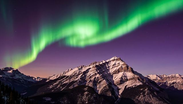 Emerald aurora dancing over snow-covered alpine peak and ridges under violet night sky