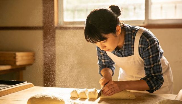 Asian baker cutting dough on wooden table, crafting artisan bread in sunlit bakery