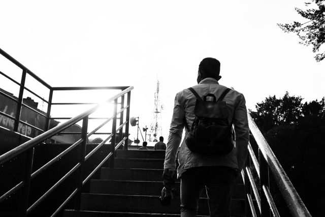 Person carrying backpack climbing outdoor steps, with an urban background and sunflare in monochrome. Useful for depicting travel, exploration, journey, solitude, and city life themes, as well as conveying movement and urban landscapes.
