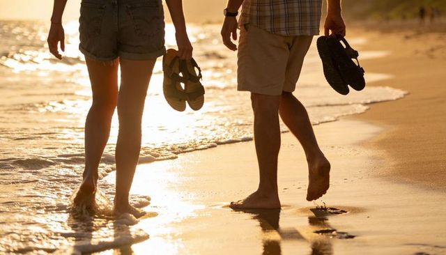Walking barefoot couple on sunlit beach at sunset carrying sandals leaving footprints