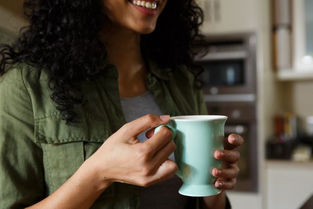 This image shows a woman with curly hair smiling while holding a green mug of coffee in a kitchen. Ideal for use in articles or advertisements related to home life, relaxation, self-care, morning routines, or lifestyle blogs.