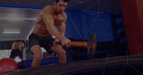 Shirtless athlete swinging sledgehammer hitting tractor tire in gym mirror workout power