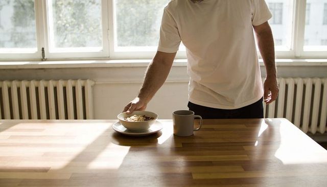 Man placing bowl of cereal on sunlit wooden kitchen table minimal morning routine