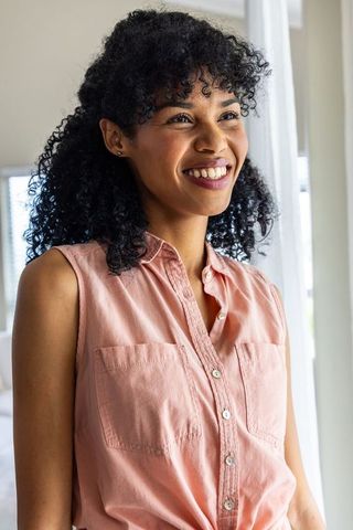 Smiling young african american woman standing by sheer-curtained window in peach blouse