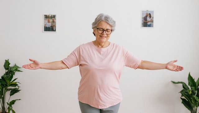 Joyful senior woman wearing glasses and pink shirt standing with arms outstretched in bright home