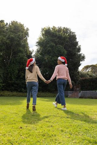 Asian female couple holding hands walking on lawn wearing santa hats and casual sweaters