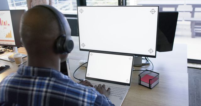 African American man wearing headset working on laptop with dual monitors in modern office. Suitable for use in business, technology, and productivity concepts, highlighting remote work, professional settings, and tech-savvy individuals.