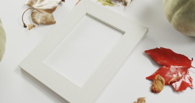 A blank white picture frame is arranged on a white surface surrounded by dried autumn leaves. This image is perfect for use in seasonal marketing, promotional materials, and decor-focused blogs. Ideal for showcasing custom artwork or photography in fall-related themes.